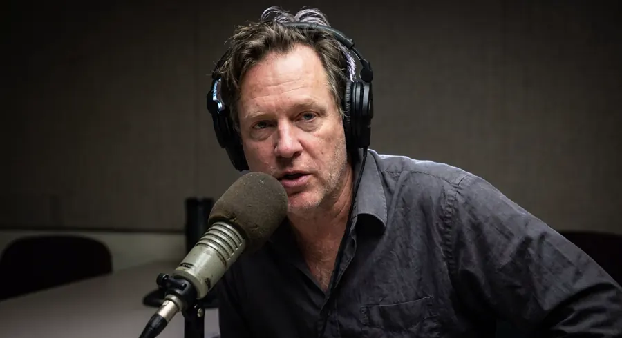 The same man at the same microphone, reshot close and lit properly, the room behind him cleared and thrown out of focus.